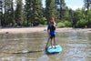 Young youth paddle boarder age 12 or 13 years old standing on his paddleboard smiling.  The photo is taken facing towards the beach from the water with amazing views of the trees and mountains in lake tahoe california.  The boy has long hair, a backwards red hat and life jacket on while he is holding his sup paddle and posing for the camera .  He is on the waterkids 8ft natural youth hardtop stand up paddle board