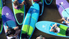 Colorful picture of a vatiety of different colors of paddle boards from a drone facing down.  All the boards are waterkids kids paddle boards with children sitting on them at a sup camp listening to their instructor teach them how to stand up paddle board.  The waterkids 8ft natural ocean blue paddleboard is in the center with a kid size adjustable paddle laying on top of it