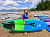 Youth athletic paddle boarder 12 to 13 years old is holding his dark blue and neon green waterkids 8ft natural hard top eps paddleboard and adjustable kids sized paddle on the beach.  Boy has a big smile and long hair, there is a pier in the background with a lot of children paying in the sand and in the water. Lake Tahoe summer fun with children playing and paddleboarding