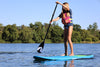 Young blond girl about 9, 10 or 11 years old is wearing a life jacket and taking a paddle stroke while she is on her blue waterkids paddle board for kids.  She is by herself in the picture while paddling on a lake with green trees behind her.  The paddle is an adjustable kids size paddle that is black with a wave logo on it