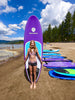 young blonde girl about 12 years old standing in the duke pose on the beach holding her stand up idle board behind her vertically while smiling and posing for the camera.  Beautiful purple color waterkids paddle board for kids and girls with children playing on the beach and in the water