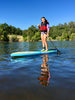 Girl with a great smile is on her waterkids 9'6 touring paddleboard tahoe SUP paddle board for kids.  The girl is on a lake and she is holding a kid size paddle, wearing a life jacket and has an ankle leash on for safety.  