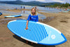 Young boy about 9 or 10 years old holds his paddleboard with a big smile while kids pay in the background in the water.  The Maui surf sup for kids is a great youth stand up paddle board that is dark and light blue with a striped traction pad for extra style