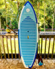 Youth surf sup paddle board made by waterkids.  Board is leaning up against a railing with palm trees in the background and a machete in a coconut ready to drink after a long day of sup surfing.  lightweight eps surf sup paddleboard for kids groms and teens. Waterkids 7'6ft Maui kids paddle board