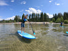 Blonde hair boy on a paddle board paddling from left to right over crystal clear water in lake tahoe california.  Colorful photo you can see the sand beneath the water, a pier, trees blue skies with puffy white clouds and mountains in the background.  Youth paddle board waterkids 9'6 ft maui surf sup